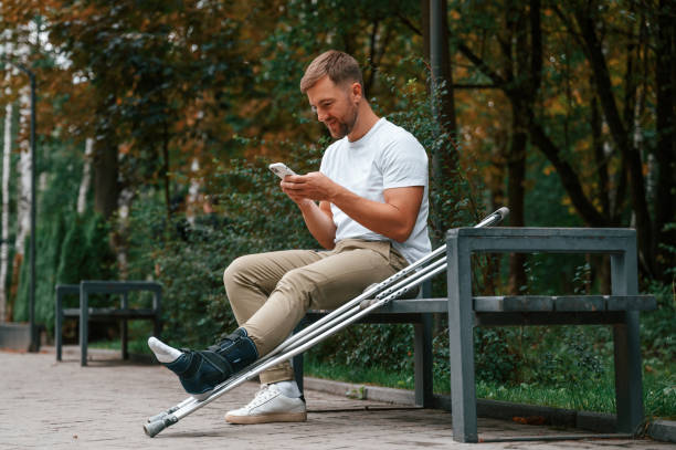 siedzenie, trzymanie smartfona. mężczyzna o kulach jest w parku na zewnątrz. uraz nogi - ludzki limb zdjęcia i obrazy z banku zdjęć