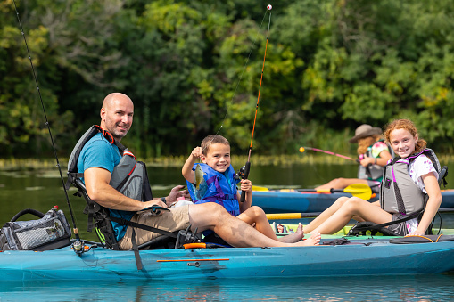 Close-up of a young boy in a kayak with his dad. The boy is holding up a bluegill sunfish he just caught while kayak fishing. His two sisters are also in this image in their own kayaks.