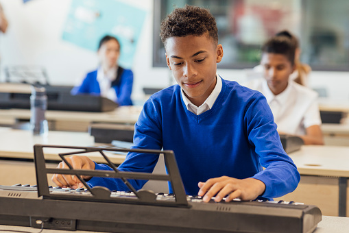 A shot of teenage male music student in a classroom learning how to play the keyboard at a school. The students are all wearing school uniforms.