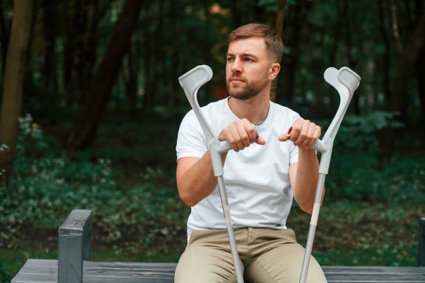 座って、退屈で悲しい気分。松葉杖をついている男が屋外の公園にいる。足をけがしている - misalignment ストックフォトと画像