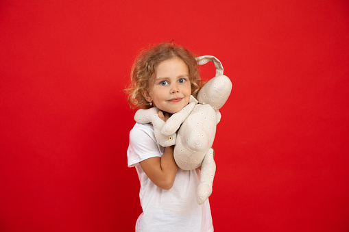 Small little expressive, positive curly blonde girl holding fluffy white rabbit toy in hands, looking at camera on red isolated studio background. Children care and protection, adoption. Copy space