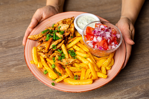 Closeup plate with fries, meat, sauce and vegetable salad isolated on wooden table background. Serving of main dish and garnish closeup. Roasted caloric food, restaurant dish, nourishing dinner.