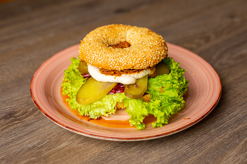 Delicious, appetizing, savory hamburger with sesame buns with hole in center on wooden table plate in restaurant. Meat, eggs burger with vegetables, salad, lettuce and cucumber on pink plate. New menu