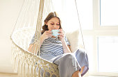 Beautiful young brunette woman drinking coffee while relaxing in knitted macrame style rocking chair