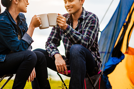 Happy Asian young couple sitting on picnic chair drinking tea and coffee while tent camp lakeside at parks outdoors on vacation holiday. Adventure lifestyle of man and woman with camping in nature.