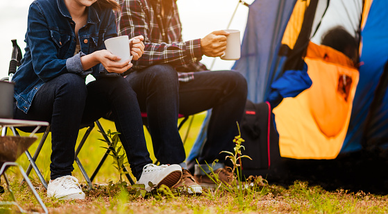 Group of Asian young sitting drinking coffee and playing guitar to happily near lake and the tent camp on outdoors camping. Lifestyle and camping concept.