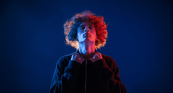 Confident look. Standing, serious facial expression. Young man with curly hair is indoors illuminated by neon lighting.