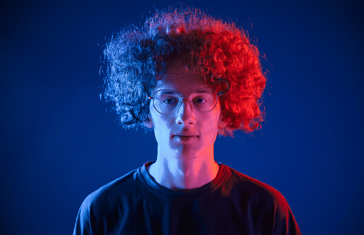 Serious facial expression. Young man with curly hair is indoors illuminated by neon lighting.