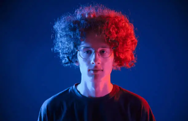 Photo of Serious facial expression. Young man with curly hair is indoors illuminated by neon lighting