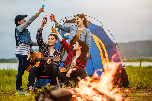 Camping tent camp in nature happy young Asian friends group in night party bonfire and the man playing guitar together girlfriend in summer at nature forest, outdoor holidays summertime concepts