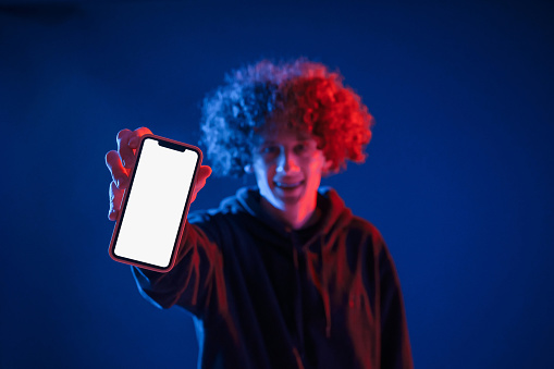 Holding smartphone and showing it to the camera. Young man with curly hair is indoors illuminated by neon lighting.