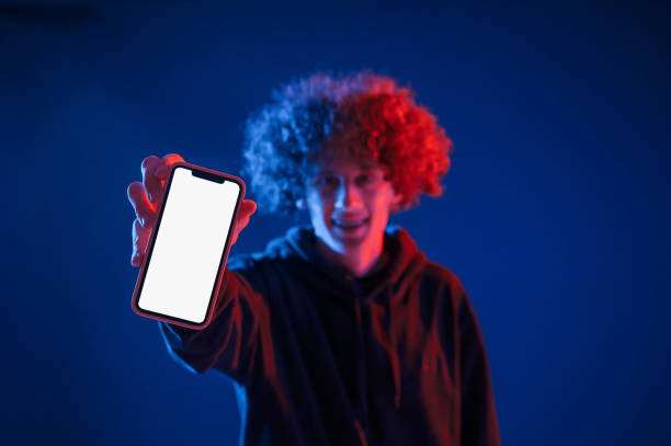 Holding smartphone and showing it to the camera. Young man with curly hair is indoors illuminated by neon lighting stock photo