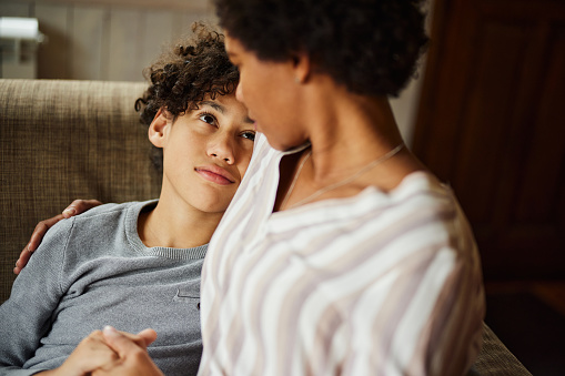 Sad African American boy talking to his single mother while feeling safe in her arms at home.