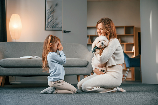 Woman is embracing the animal. Mother with daughter is at home with maltese dog.