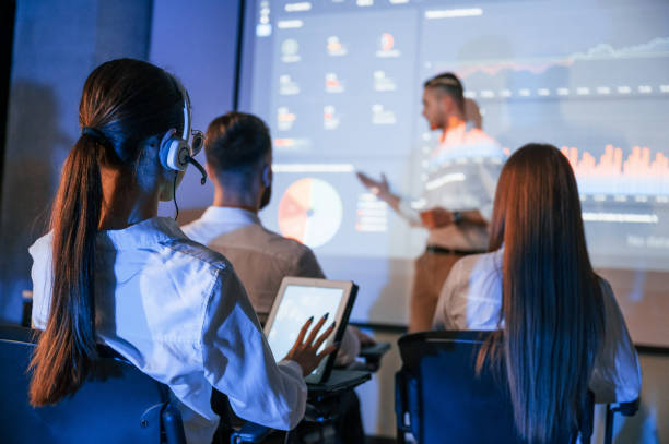 istock Master class. Male leader talking to employees, showing the plan on the projector in office of stock exchange company 1457992592