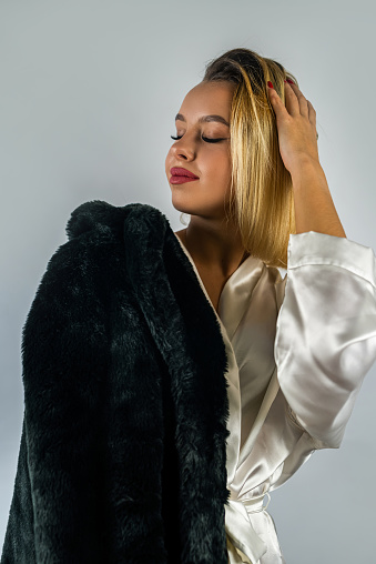 young beautiful blonde in a white silk robe with a fur coat on top. isolated on plain background. concept of heat