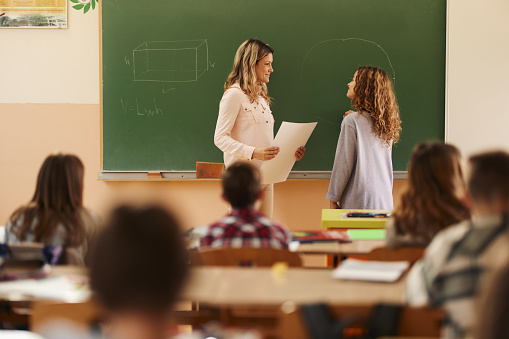 Smiling female teacher talking to schoolgirl in front of blackboard during a mathematics class at elementary school.