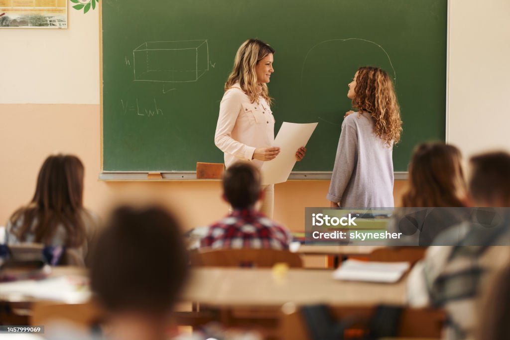 Learning geometry in elementary school! Smiling female teacher talking to schoolgirl in front of blackboard during a mathematics class at elementary school. Classroom Stock Photo