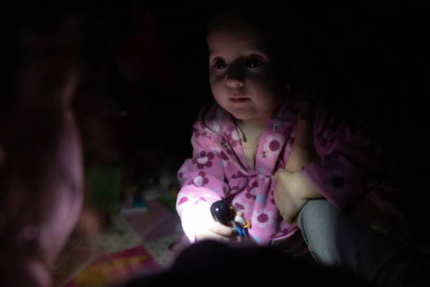 The mother plays with her daughter during a blackout. They are sitting on the floor with a flashlight in complete darkness due to a blackout.