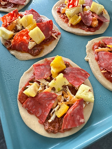 Stock photo showing close-up, elevated view of uncooked mini pizzas filled with sliced pepperoni, pineapple chunks and grated cheese prepared on a baking sheet, ready to put in a hot oven