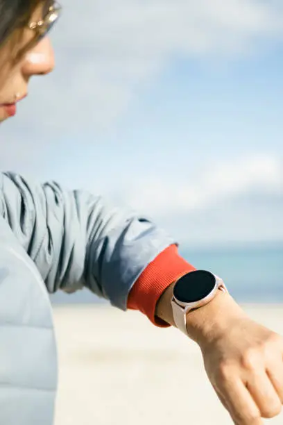 Photo of Waist up shot of happy brunette woman dressed in anorak checks fitness results on smartwatch poses against the beach on the coast. Healthy lifestyle