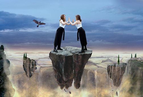 Composite image of same woman standing face to face leaning on her own hands, flying on the huge rock where the total cataclysm is happening, the land is cracking, debris, dust, fire, rocks with trees and houses are falling apart.\nThe image presents: supporting yourself in difficult times, lean into your own power, believe, depend, lean on yourself, be yourself bridge over troubled water, we are our own best therapists.