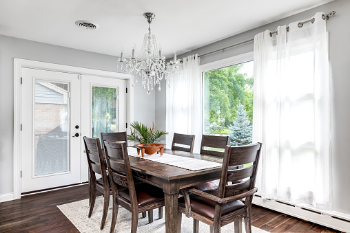 A large dining room table with a fancy chandelier hanging from above. The windows and door show beautiful green trees.