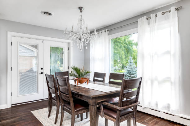 A dining room with a chandelier hanging above the wood table. A large dining room table with a fancy chandelier hanging from above. The windows and door show beautiful green trees. dining room stock pictures, royalty-free photos & images