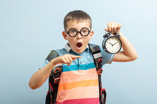A cute school boy in glasses with an alarm clock against blue background