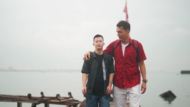 Asian Chinese down syndrome young man walking with father on wooden bridge Penang seaside during overcast morning