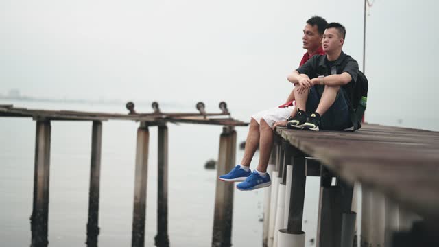 Asian Chinese down syndrome young man sitting side by side with father on wooden bridge seaside during overcast morning