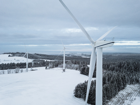 Wind Turbines in white snow covered rural Winter Landscape under partly sunny dramatic winter skyscape. Green Renewable Energy, Alternative Energy Environment Concept Shot. Drone Point of View. Baden Württemberg, Germany, Europe