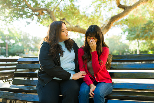 Worried young woman comforting and hugging her sad best friend crying while taking at the park