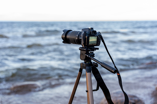 a camera on a tripod mounted on the seashore in the process of shooting waves in cold weather. the horizon line is visible, the beautiful sky and the bay. photographer takes scenic views. travel, discovery and photo