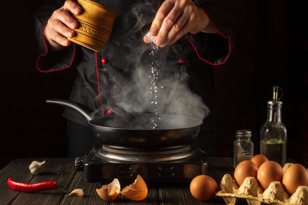 istock Professional chef adds salt while cooking eggs or an omelet in pan. Work environment on kitchen table. Menu idea for a restaurant or hotel. 1458044282