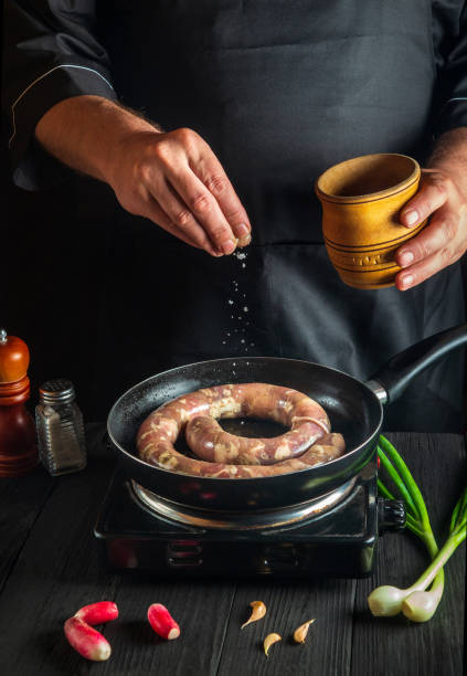 The chef adds salt to pan with raw meat sausage. Preparation for cooking sausages in the kitchen of a restaurant or cafe on table with vegetables and spices