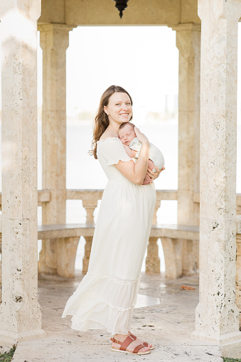Lifestyle Newborn Family Photo Palm Beach, Florida in November of 2022. 
A Young Mother Holding Her Newborn Son Relaxed & Enjoying QualityTime Snuggling Her Boy While in a Stone Gazebo on the Water in a Relaxing Meditation Garden in Palm Beach, Florida for Lifestyle Newborn Family Photos.