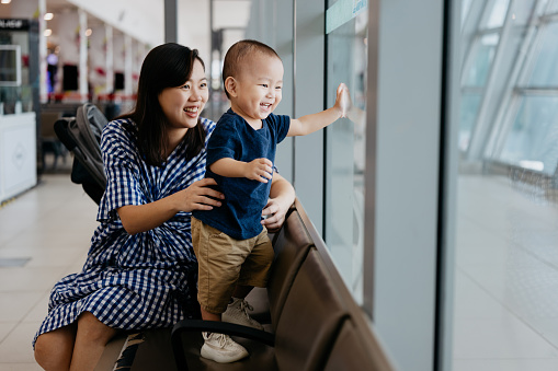 Image of an Asian Chinese woman and her baby boy looking out window for airplane at airport terminal