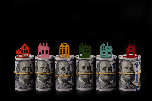 Stacks of American dollars and miniature multi-colored houses standing on them on a black background