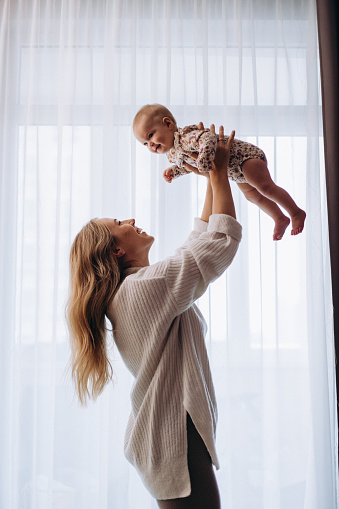 A loving beautiful mother lifts her child up to admire her near the window at home