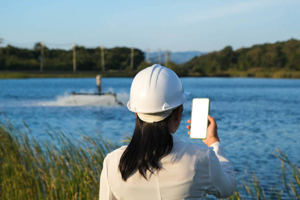 istock Environmental engineer wearing a white helmet uses a mobile phone to operate an oxygen turbine aerator in a pond. Water and ecology concept. 1458142746
