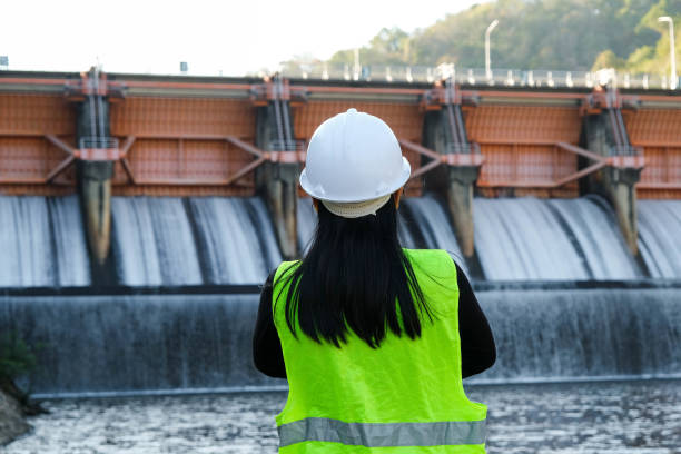 녹색 조끼와 헬멧을 쓴 여성 엔지니어의 뒷모습은 수력 발전소와 관개가 있는 댐의 배경에 밖에 서 있습니다. - dam 뉴스 사진 이미지