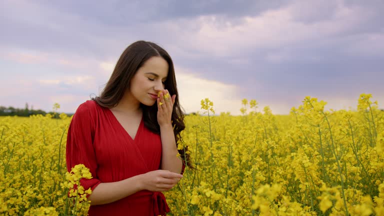 Medium, slow motion shot of brunette in red dress, surrounded by yellow snapdragon flowers, smelling one of them