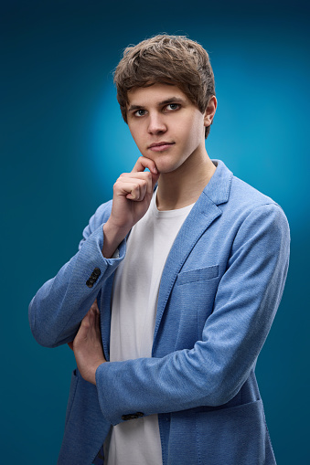 handsome thoughtful caucasian man holding arms folded, touching chin with hand and looking at camera on blue background