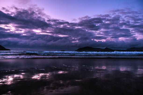Dramatic light breaks through storm clouds over a vast, wet beach at low tide, creating a moody, atmospheric scene.
