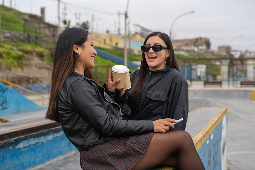 Portrait of two latin young women drinking take away coffee sitting on an urban park