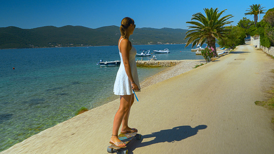 Female tourist is riding an elongboard while exploring Peljesac during her vacation. Young Caucasian woman in a white sundress rides an electric longboard along a scenic coastal road in Croatia.