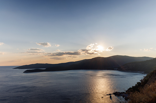 Bright dazzling golden summer sun in the blue cloudy evening sky illuminates all the silhouettes of the peaks of the Balkan Montenegrin mountains and the coast of the famous touristic Kotor Bay
