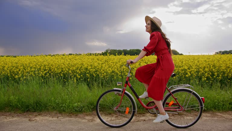 Tracking, side view of a brunette female cycling through a field of vibrant yellow snapdragon flowers, wearing a red dress and a beige hat