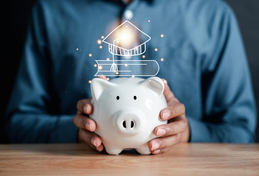 Businessman holding coin into piggy bank with graduation cap and book icons. saving money for future education. Education fees and cost of college graduate. Tuition fees and total cost in education.